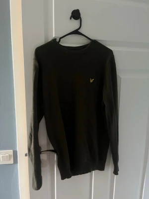 Mörkgrön tröja från Lyle & Scott - Mörkgrön långärmad tröja från Lyle & Scott med rund halsringning och den klassiska gula loggan broderad på bröstet. Tröjan har en enkel och stilren design, perfekt för en avslappnad look.