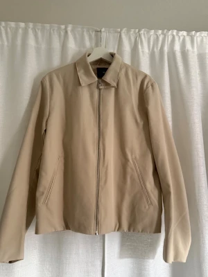 Beige minimalistisk jacka med dragkedja - Stilren beige jacka med rak passform och klassisk krage. Jackan har dragkedja framtill och två snedställda fickor. Perfekt för dig som gillar en clean och enkel look. 