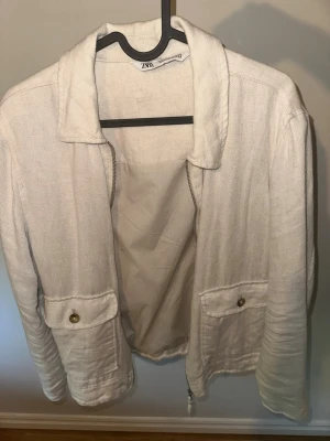 Beige jacka från Zara i linne - Stilren beige jacka från Zara i linne med klassisk krage, två fickor framtill med knappar och dragkedja. Perfekt för dig som gillar en enkel och clean look. PERFEKT FÖR SOMMAREN. Storlek S men ganska liten i storleken.