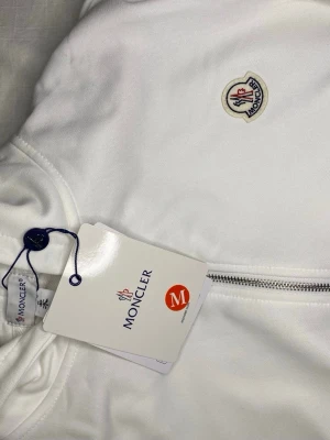 Vit hoodie med dragkedja från Moncler - Snygg vit hoodie från Moncler med dragkedja framtill och klassisk logga på bröstet. Tillverkad i mjukt material som känns skönt mot huden. Perfekt för dig som gillar stilrena och exklusiva plagg. Långärmad och med ribbade muddar.