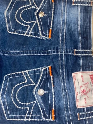 Blå True Religion jeans med kontrastsömmar - Säljer ett par blå jeans från True Religion med ikoniska kontrastsömmar och detaljerade bakfickor med vita och orange sömmar. Klassisk femficksmodell och silverfärgade knappar. Perfekta för dig som vill sticka ut med en streetig look.