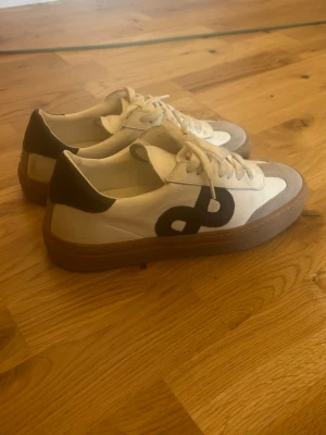 Pavement KOHIA vita sneakers med svart detalj - Snygga vita sneakers från pavement KOHIA med svart logga på sidan, beige mockadetaljer och bruna gummisulor. Skorna har lågt skaft och vita snören. Perfekta för dig som gillar stilrena och trendiga sneakers med en unik touch. Dom är knappt använda.