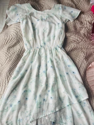 Kappahl kortklänning - Söt ljus kortklänning med blommigt mönster i pastellfärger. Klänningen har korta ärmar, rund halsringning och elastisk midja. Detalj med knapp i nacken och omlottliknande kjol. Perfekt för sommarens alla tillfällen!