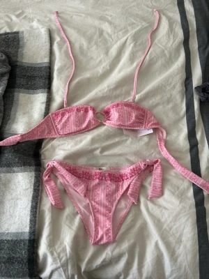 Rosa mönstrad bikini från Gina Tricot - Söt bikini från Gina Tricot i rosa med ljusa och mörkare rosa ränder och blommönster. Överdelen har knytband och en dekorativ ring i mitten, underdelen har knyt i sidorna. Perfekt för stranden eller poolen! Aldrig andvänd 