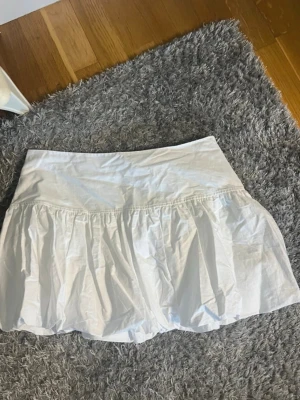 Vit kortkjol från Weekday - Säljer en vit kortkjol från Weekday i luftig bomull. Kjolen har en volangig design och bred midja som ger en snygg siluett. Perfekt för dig som gillar en enkel men trendig look.