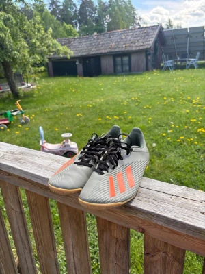 Adidas grå-orange fotbollsskor - Snygga fotbollsskor från Adidas i grått med orangea stripes på sidan och svart snörning. Skorna har låg profil och gummisula som passar perfekt för inomhusspel. Tillverkade i syntetmaterial för bra komfort och kontroll på planen.