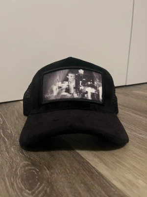 Svart Divirtido keps med tryck - Svart truckerkeps från Divirtido med mesh på sidorna och bak och frotté som material. Framtill finns en svartvit patch med ett tryck av ett retro foto. Böjd skärm och justerbar passform. Perfekt för en streetwear-look. Pris kan absolut diskuteras.