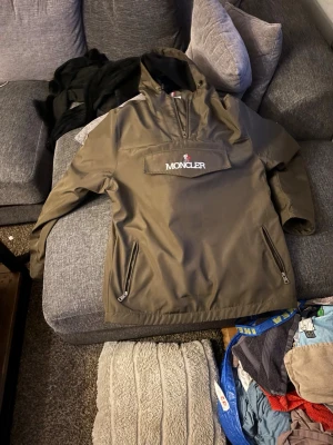 Olivgrön Moncler vindjacka med dragkedja - Snygg olivgrön vindjacka från Moncler med halv dragkedja framtill och stor ficka med broderad logga på bröstet. Jackan har två snedställda fickor med dragkedja och huva. Perfekt för blåsiga dagar och streetwear-stil.