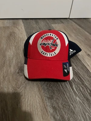 Washington Capitals keps från Adidas - Snygg keps från Adidas med Washington Capitals-logga framtill. Kepsen är röd med vita och mörkblå detaljer samt broderad logotyp. Perfekt för dig som gillar hockey och vill visa vilket lag du hejar på.  Pris kan absolut diskuteras.   