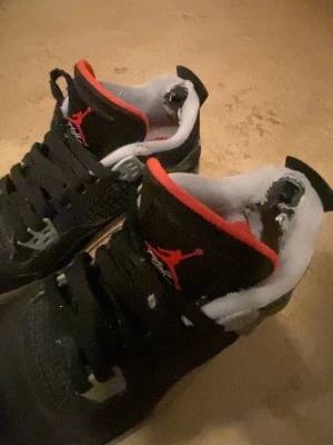 Svarta Air Jordan 4 Retro sneakers - PRIS KAN DISKUTERAS! Grymma Air Jordan 4 Retro sneakers i svart med röda och grå detaljer. Klassisk siluett med meshpaneler, plastdetaljer och synlig Air-sula. Tungan har röd Jumpman-logga och insidan är vit. Perfekta för dig som gillar streetwear och ikoniska sneakers. Det är storlek 40 men kan passa som 39.