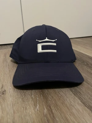 Mörkblå Cobra keps med vit logga - Snygg mörkblå keps med böjd skärm och vit broderad logga framtill som föreställer ett C med en krona ovanför. Klassisk sexpanelsdesign och justerbar baksida. Perfekt accessoar för en sportig och trendig look. Pris kan absolut diskuteras.   