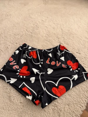 Svarta shorts med hjärtan - Coola svarta shorts med röda och vita hjärtan samt texten 'Love' tryckt över hela plagget. De har elastisk midja och är perfekta för en avslappnad stil. Materialet känns mjukt och lätt.