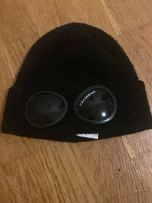 Svart mössa från C.P. Company - Svart ribbstickad mössa från C.P. Company med ikoniska runda glasögon-linser framtill på uppviket. Klassisk beanie-modell i mjukt material som sitter skönt på huvudet. Perfekt för dig som vill sticka ut med en streetwear-inspirerad stil.