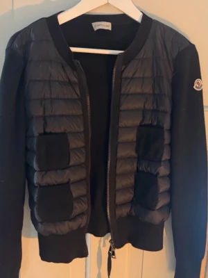 Moncler cardigan  - Snygg svart cardiganjacka från Moncler med quiltad front och mjuka stickade ärmar. Jackan har dragkedja, ribbade muddar och två fickor framtill. Ikonisk Moncler-logga på ärmen. Perfekt för lager på lager och streetstyle.