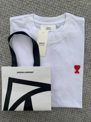 Vit t-shirt från Ami med rött hjärta - Vit t-shirt från Ami Alexandre Mattiussi med klassisk passform och rund hals. På bröstet finns Ami:s ikoniska röda hjärta-broderi. Tillverkad i mjuk bomull och perfekt för en clean och stilren look.