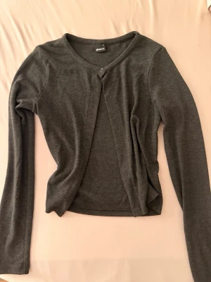 Mörkgrå tunn kofta från Gina Tricot - Snygg mörkgrå tunn kofta från Gina Tricot med rund halsringning och en knapp framtill. Långärmad och i mjukt, stretchigt material som passar perfekt till lager-på-lager-stil. Storlek S