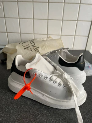 Alexander McQueen vita sneakers - Stilrena vita sneakers från Alexander McQueen med svart hälparti och guldfärgad logga. Skorna har tjock sula, breda vita skosnören och detaljer i läder. Perfekta för dig som gillar en clean och trendig look.