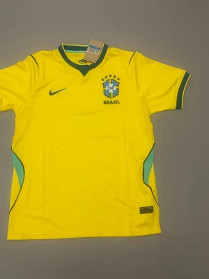 Brasiliens vm tröja - Gul Brasilien fotbollströja från Nike med gröna detaljer och CBF-emblem på bröstet. Tröjan har korta ärmar, V-ringad krage med grönt kantband och är tillverkad i lätt och ventilerande material. Perfekt för fotbollsträning eller att bära som supporter.