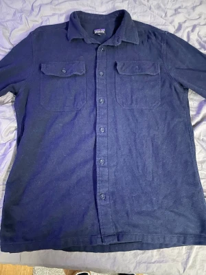 Mörkblå overshirt från Patagonia - Stilren mörkblå overshirt från Patagonia med klassisk krage och två bröstfickor med lock och knapp. Skjortan har knäppning hela vägen och är tillverkad i ett mjukt, lite tjockare tyg som passar perfekt som lager. Enkel och tidlös design.