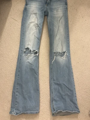 Ljusblå bootcut jeans från Nelly - Snygga ljusblå bootcut jeans från Nelly med slitna detaljer och stora hål vid knäna. Jeansen har klassisk femficksdesign, bälteshällor och dragkedjegylf. Perfekta för en avslappnad och trendig look.