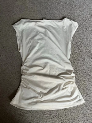Vit ärmlös topp med rynkade sidor - Snygg vit topp med ärmlös design och rynkade detaljer på sidorna. Toppens enkla snitt gör den lätt att matcha och den har en mjuk, stretchig känsla. Perfekt för dig som gillar stilrena plagg med en twist.