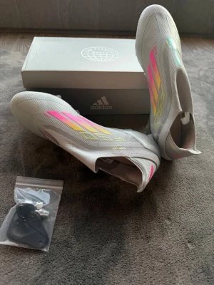 Adidas F50 elite SG - Snygga Adidas F50+ fotbollsskor i silver med neonrosa och gula detaljer. Skorna har en strömlinjeformad design utan snörning och metall-dobbar för bästa grepp på planen. Ovandelen är i lätt syntetmaterial och sulan har inslag av neonfärg. Perfekta för dig som vill sticka ut på matchen. Inte använda!!