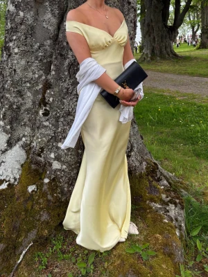 Ljusgul offshoulder långklänning satin - Jättefin och snygg klänning, passar till bal eller likande! Ljusgul långklänning i satin med offshoulder-skärning och lätt v-ringad byst. Den är öppen i ryggen. En söm vid sidan är uppsrättad, inget som syns men gjordes för att inte sitta åt så hårt längs sidan.