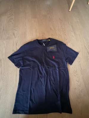 Mörkblå samt svart t- shirt - Snygg mörkblå t-shirt från P R L med klassisk rund hals och röd broderad logga på bröstet. Tillverkad i mjuk bomull och har en normal passform. Perfekt för en stilren och avslappnad look. OBS endas 1 T-shirt till priset