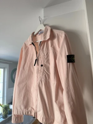 Ljusrosa overshirt från Stone Island - Säljer en ljusrosa overshirt från Stone Island med klassisk krage, dragkedja och knappstängning framtill. Har en bröstficka och den ikoniska loggan på vänster ärm. Perfekt för lager-på-lager och streetwear-stil.