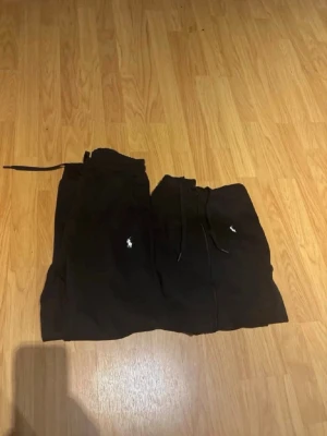 Svart loungewear-set från Polo Ralph Lauren - Svart loungewear-set från Polo Ralph Lauren med matchande hoodie och byxor. Setet har den klassiska vita Polo-loggan broderad på både tröjan och byxorna. Hoodien har dragkedja och justerbar huva, byxorna har dragsko i midjan. Perfekt för chill dagar hemma. 