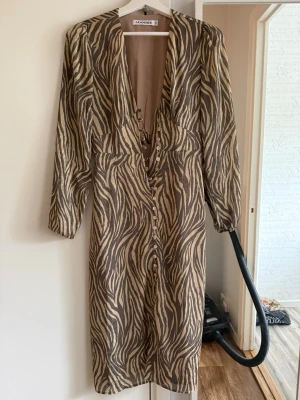 Adoor - Säljer en zebramönstrad klänning från Adoore i beige och brunt. Klänningen har v-ringning, knappar framtill och långa ärmar. Snygg passform och trendigt mönster som sticker ut.