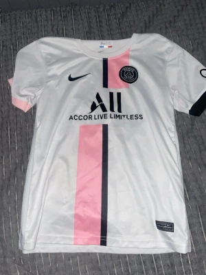 PSG vit och rosa matchtröja Nike - Säljer en vit Paris Saint-Germain matchtröja från Nike med rosa och svart vertikal rand, PSG-logga och sponsortryck på bröstet.Perfekt för fotbollsträning eller att bära som supporter.