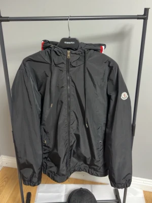 Svart vindjacka från Moncler - Snygg svart vindjacka från Moncler med huva och dragkedja framtill. Jackan har dragsko i huvan, två fickor med dragkedja och Moncler-logga på ärmen. Materialet är glansigt och lätt, perfekt för blåsiga dagar. Röd och vit detalj på huvan ger extra stil.              Priset kan diskuteras vid snabb affär!