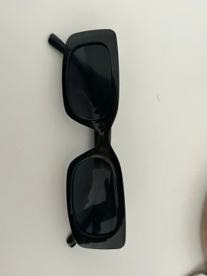 Svarta rektangulära solglasögon - Snygga svarta solglasögon med rektangulära bågar och mörka glas. De har en trendig, chunky design som passar perfekt till en modern stil. Bågarna är i plast och ger en cool vibe till vilken outfit som helst.