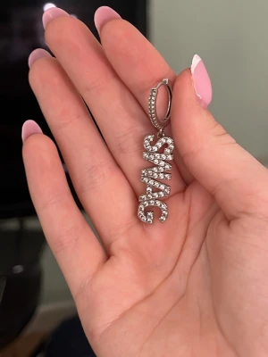 Ioaku örhänge  - Coolt örhänge i silverfärg med ordet SWAG i stora bokstäver täckta av glittrande stenar. Hängande modell med små pärlliknande detaljer på både ringen och bokstäverna. Perfekt accessoar för att sticka ut och visa attityd.