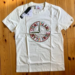 Vit Stone Island x Supreme t-shirt - Vit t-shirt från Stone Island i samarbete med Supreme. Stor broderad kompasslogga i flera färger på bröstet. Klassisk passform med rund hals och korta ärmar. Perfekt för dig som gillar streetwear och unika samarbeten.