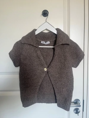 Brun stickad kofta från Zara - Kortärmad brun stickad kofta från Zara med bred krage och en dekorativ knapp framtill. Koftan har en avslappnad passform och ribbade muddar vid ärmslut och nederkant.  Inga defekter 💗