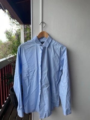 Ljusblå skjorta från Selected Homme - Klassisk ljusblå skjorta från Selected Homme med knappar framtill och lång ärm. Skjortan har en stilren krage och är tillverkad i bomull, perfekt för en clean och enkel look. Slim fit.
