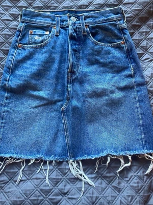 Levi's jeanskjol med fransad kant - Snygg jeanskjol från Levi's i klassisk blå denim. Kjolen har fem fickor, fransad nederkant och slitna detaljer på bakfickorna. Perfekt för dig som gillar en avslappnad och trendig stil.