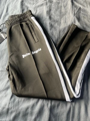 Palm Angels olivgröna mjukisbyxor - Olivgröna mjukisbyxor från Palm Angels med vita sidoränder och logga i vitt framtill. Byxorna har resår i midjan och raka ben. Snygg streetwear-look med bekväm passform, perfekta för en avslappnad stil.