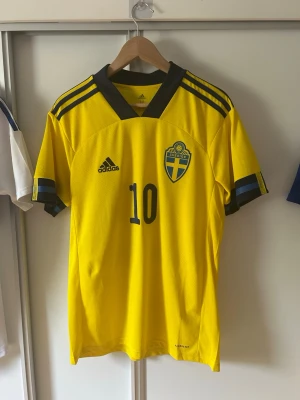 Sverige matchtröja Forsberg #10 Adidas - Gul Sverige matchtröja från Adidas med mörkblå detaljer, nummer 10 och Forsberg på ryggen. Tröjan har officiellt landslagsmärke på bröstet, V-ringad krage och korta ärmar med blå ränder. Perfekt för fotbollsfans och samlare.