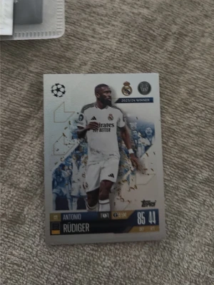 Samlarbild Real Madrid Antonio Rüdiger - Samlarbild från Topps med Antonio Rüdiger i Real Madrids vita matchställ. Kortet har detaljer i blått, guld och svart samt Champions League-logga och klubbmärke. Perfekt för dig som gillar fotboll och samlarkort.