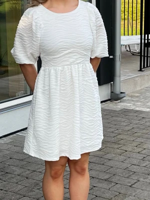 Vit kort klänning med puffärm - Superfin vit kort klänning med puffärmar och strukturerat tyg i vågigt mönster. Klänningen har rund halsringning och markerad midja för en söt och trendig look. Perfekt för sommarens alla tillfällen!