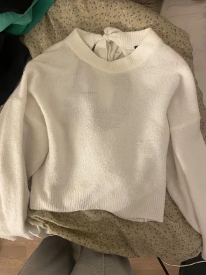 Vit stickad tröja med rosett i ryggen - Säljer en vit stickad tröja med rund hals och långa ärmar. Tröjan har en snygg cut-out i ryggen med en bred rosett som knyts upptill. Mjuk och skön kvalitet.
