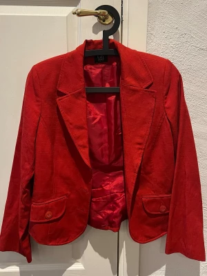 GR DESIGN röd kavaj, storlek 38 - Röd kavaj med krage och slag. Ärmarna är långa och kavajen har en figurnära passform med sömmar som framhäver midjan. Baksidan har en slät design med diskreta sömmar.