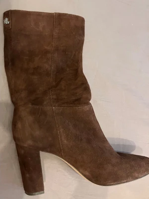 Bruna mockastövlar från Ralph Lauren - Snygga bruna stövlar i mocka från Ralph Lauren med bred klack och något spetsig tå. Stövlarna har ett diskret RLL-emblem upptill och en slouchy passform som ger en avslappnad men trendig look. Storlek 38 men passar 39