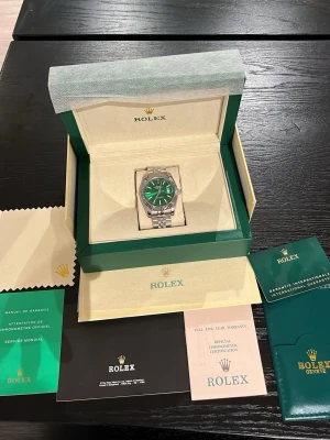 Rolex klocka med grön urtavla - Exklusiv Rolex klocka med grön urtavla och silverfärgat armband i rostfritt stål. Klockan har klassisk rund form, räfflad bezel och tydliga indexmarkeringar. Levereras i originalbox med certifikat och tillbehör.
