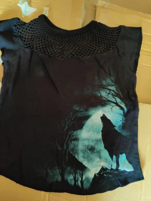 Svart topp med nät och vargmotiv goth  - Cool svart topp med nät över axlarna och ett tryck av en ylande varg mot en blåmålad måne. Toppen har korta ärmar och en rund halsringning. Perfekt för dig som gillar unika och lite alternativa plagg.