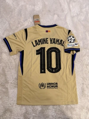 FC Barcelona Lamine Yamal tröja - Snygg beige FC Barcelona matchtröja med Lamine Yamal och nummer 10 på ryggen. Tröjan har svarta detaljer, klubbmärke, Champions League-märke på ärmen och UNHCR-logga. Tillverkad i funktionsmaterial, perfekt för fotbollsträning eller match.