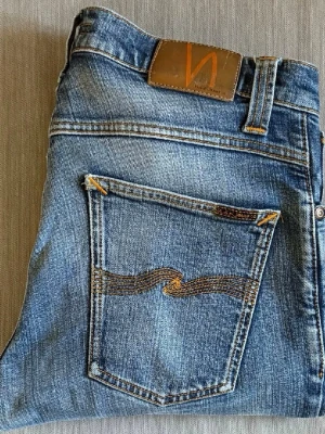 Blå jeans från Nudie Jeans - Säljer ett par klassiska blå jeans från Nudie Jeans med snygga slitningar och kontrastsömmar. Jeansen har fem fickor, normal midja och Nudie Jeans ikoniska broderi på bakfickan. Tillverkade i denim av bomull. De är grim tim w32 l32